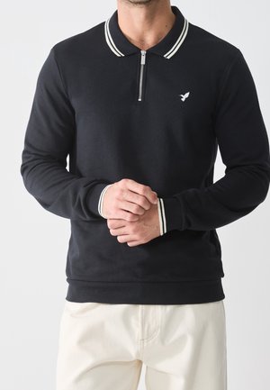 Hombre vistiendo una camisa tipo polo de manga larga negra con ribetes blancos en el cuello y los puños, cremallera frontal y pequeño logotipo de pájaro en el pecho.