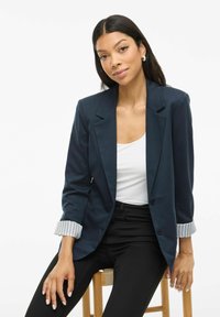 Femeie cu păr lung și închis la culoare, purtând cercei cerc, un sacou bleumarin cu manșete striped rulate, o bluză albă și pantaloni negri, stând pe un scaun din lemn.