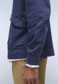 Φούτερ σε ναυτικό μπλε χρώμα με μακριά μανίκια, διακοσμημένο με τσέπη και ανάγλυφη λεπτομέρεια λογότυπου. Φορεμένο πάνω από λευκή μπλούζα, σε συνδυασμό με χακί παντελόνι.