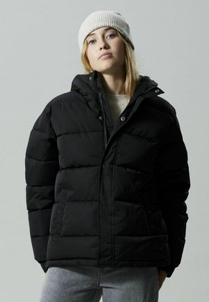 Schwarze Pufferjacke mit Kapuze, ausgestattet mit horizontaler Steppung, Druckknopfverschluss und Seitentaschen; über einem leichten Pullover getragen.