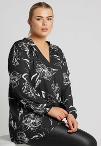 Blusa nera con motivo floreale bianco, scollatura a V e maniche lunghe con polsini arricciati. Tessuto morbido, vestibilità rilassata. Abbinata a leggings neri.