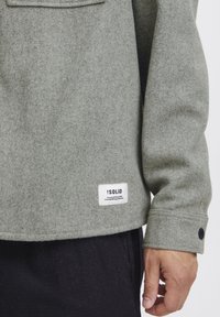 Grijze wolmix shirt met een borstzak, knoopmanchetten en een merketiket aan de onderrand. Zachte textuur en casual ontwerp.