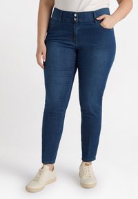 Pantalones skiny de mezclilla azul oscuro con cintura alta, dos bolsillos frontales y cierre de doble botón. Combinados con zapatillas crema. Textura de mezclilla suave.
