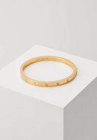 Das goldene Armreifarmband zeichnet sich durch ein glattes Design aus, das mit mehreren klaren Edelsteinen entlang seines Umfangs verziert ist. Innen ist "kate spade NEW YORK" eingraviert.