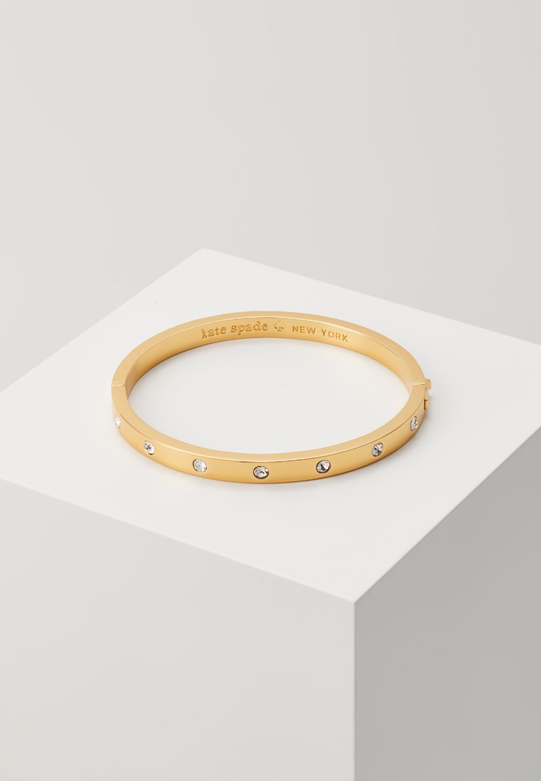 Das goldene Armreifarmband zeichnet sich durch ein glattes Design aus, das mit mehreren klaren Edelsteinen entlang seines Umfangs verziert ist. Innen ist "kate spade NEW YORK" eingraviert.