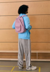 Ροζ σακίδιο με σχέδια και μπροστινή τσέπη, φορεμένο πάνω από ανοιχτό μπλε μπουφάν και γκρι παντελόνι από κορδόνι. Ξύλινος τοίχος στο φόντο.