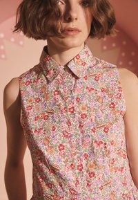 Cvetlična majica brez rokavov z ovratnikom. Ima večbarven vzorec cvetov in listov na svetlo roza ozadju.