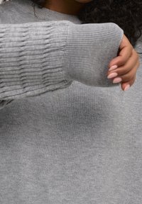 Woman wearing a ribbed gray sweater with extended sleeves covering part of her hand and curled nails visible.