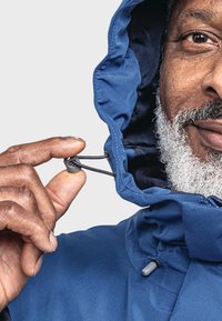 Blaues Kapuzenjackett mit strukturiertem Finish; eine Hand zieht den Kordelzug am Kragen, wodurch ein dicker grauer Bart sichtbar wird.