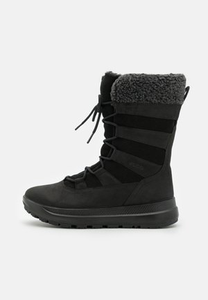 Svart vanntett støvel med semsket ytre, fleece-fôret krage og elastiske snører. Har en teksturert gummisåle for grep og støtte.
