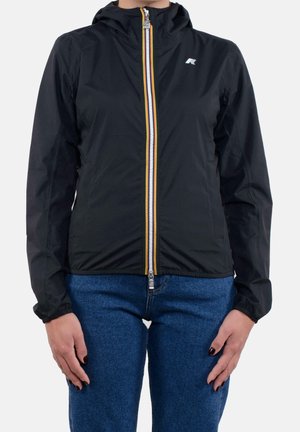 Kvinde iført en sort hættetrøje med en flerfarvet stribet lynlås og blå jeans, stående med hænderne afslappet ved siden af kroppen.