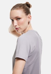 Camiseta deportiva gris con un patrón texturizado, de manga corta y cuello redondo, presentada en un modelo con el cabello recogido.
