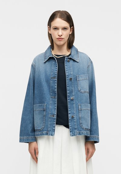 Femme portant une veste en jean bleu clair sur un haut noir et une jupe blanche, accessoirisée d'un collier de perles, devant un fond uni.