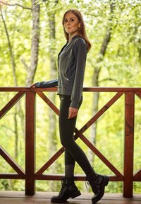 Grå zip-up jakke med to forlommer, parret med sorte leggings og sorte snørebåndsstøvler, udendørs med en baggrund af trægelænder.