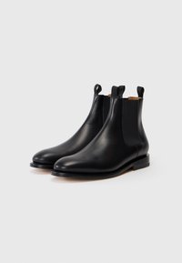 O pereche de bote din piele neagră, elegante, tip Chelsea, cu panouri laterale elastice și curele de tragere, plasate pe un fundal alb simplu.