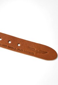 Bouton de ceinture en cuir brun avec trois trous, texte en relief "P.ME LEGEND" et un emblème ailé sur un fond blanc.