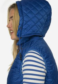 Modrý prošívaný vestový kabát s kapucí, který má elastickou šňůrku a kontrastní žlutou podšívku. Nositý přes bílé a modré pruhované tričko.