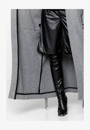 Długi szary płaszcz z czarnymi akcentami, wykonany z teksturowanej tkaniny. Pod płaszczem czarne skórzane spodnie z bocznym rozcięciem i czarne botki do kolan.