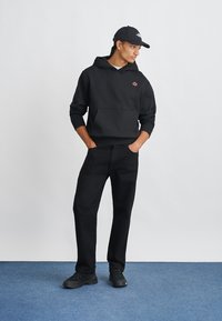 Sweatshirt preto com capuz e bolso frontal, combinado com calças pretas e ténis pretos. O modelo está de pé sobre um tapete azul contra um fundo simples.