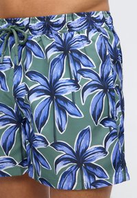 Ανδρικά μαγιό σορτς με μπλε τροπικό σχέδιο φοίνικα και πράσινο κορδόνι στη μέση, σε ανοιχτό πράσινο φόντο.