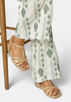 Pies con sandalias de cuña beige y pantalones crema con bordado floral verde, descansando sobre la pata de una silla de madera sobre un fondo blanco.