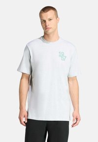 Vaaleanharmaa puuvillainen t-paita, jossa on lyhyet hihat, pyöreä kaula-aukko ja mintunvihreä graafinen kuvio, jossa lukee "1957" vasemmalla rintapuolella.