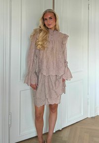 Plavuša stoji u zatvorenom prostoru noseći bež čipkastu haljinu s dugim rukavima, volanima i valovitim rubovima, ispred bijelih panela vrata.