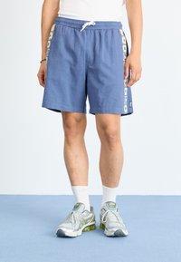 Lys blå shorts med elastisk midjebånd og snøring. Sidepanelet har et blomsterdesign. Båret med grå sportsko og hvite sokker.