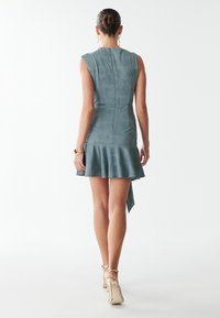 Short dress in textured teal fabric, featuring a fitted bodice, flared hem, and asymmetric back detail. Gold-tone hardware accents.