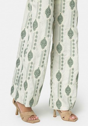 Pantalones blancos de pierna ancha con patrones de hojas bordadas en verde, combinados con sandalias de tacón alto abiertas en ante beige.