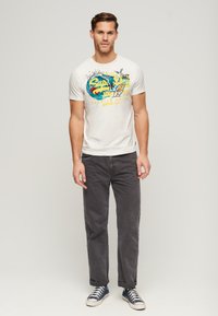 Camiseta de algodón blanca con un diseño gráfico colorido de olas y una tabla de surf, combinada con pantalones gris oscuro y zapatillas azules.