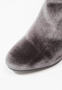 Botín de terciopelo gris con un acabado suave, punta afilada y un patrón ligeramente texturizado. Suela de goma negra y forma cónica.