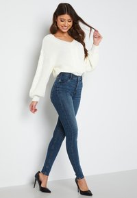 Bílý pletený svetr s výstřihem do V, kombinovaný s tmavě modrými skinny džíny a černými vysokými podpatky, zdůrazňuje přiléhavý siluetu.