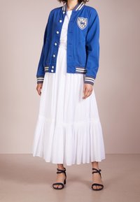 Modrá baseballová bunda s bílými knoflíky a pruhovanými manžetami, nošená přes dlouhé, bílé, mnohoúrovňové šaty. Outfit doplňují námořnické sandály.