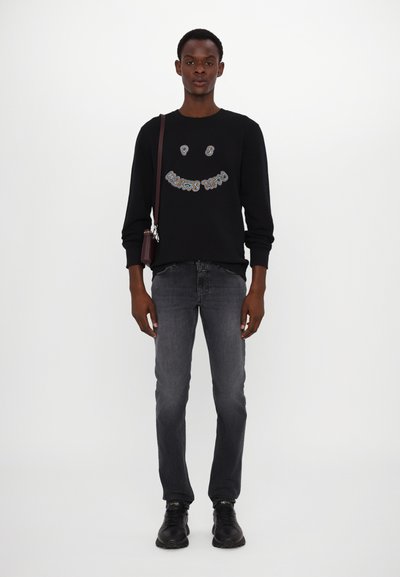 Sudadera negra con diseño de cara sonriente, jeans grises y zapatos negros. Presenta un bolso de hombro y un ajuste relajado. Textura suave.