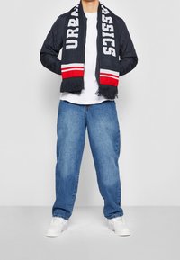 Mörkblå quiltad bomberjacka, vit t-shirt, blå baggy jeans, vita sneakers; mörkblå scarves med "URBAN CLASSICS"-text och röda accenter, fransade kanter.