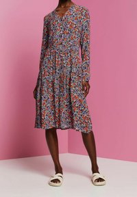 Nainen, joka on pukeutunut polvenmittaiseen, pitkähihamaiseen kukkamekkoon, jossa on oransseja, violetteja ja valkoisia kukkia, seisoo vaaleanpunaisen seinän edessä valkoisissa sandaaleissa.