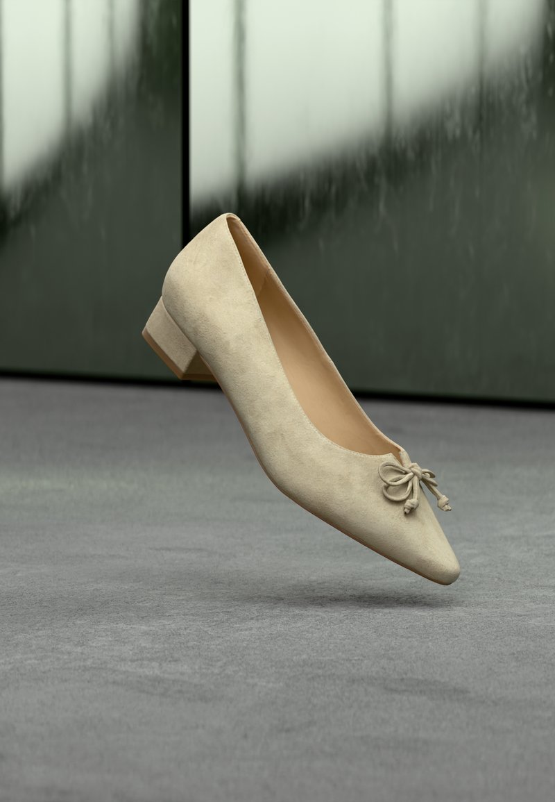 Chaussure pour femme en daim beige à petit talon avec un petit nœud décoratif flottant au-dessus d'une surface grise sur un fond vert flou.