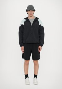 Črna vetrovka z svetlo modrimi detajli, zadrgo spredaj in kapuco. Črne kratke hlače in siva kapa dopolnjujeta videz. Bele tene z vezalkami.