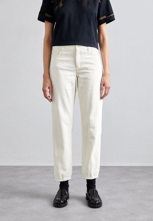 Mujer vistiendo vaqueros blancos de tiro alto y pierna recta, top negro corto con mangas de encaje, calcetines negros y mocasines negros, de pie sobre un suelo gris.