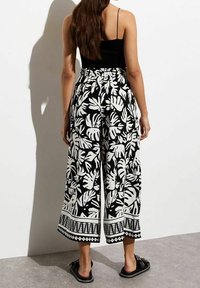 Femme portant un haut noir sans manches et un pantalon large taille haute avec un imprimé feuilles noir et blanc, debout contre un mur blanc.