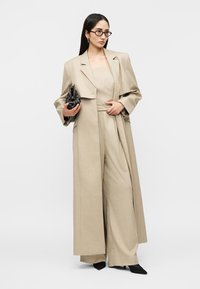 Γυναίκα που φοράει μπεζ παντελόνια με φαρδύ πόδι, ταιριαστό κορμάκι χωρίς τιράντες, μακρύ παλτό, μαύρα μυτερά παπούτσια, γυαλιά, κρατώντας μαύρο τσαντάκι με τρουκς.