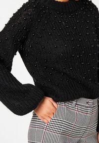 Pull en maille noire avec des détails en perles 3D sur le devant, manches longues et encolure ronde, associé à un pantalon à carreaux.