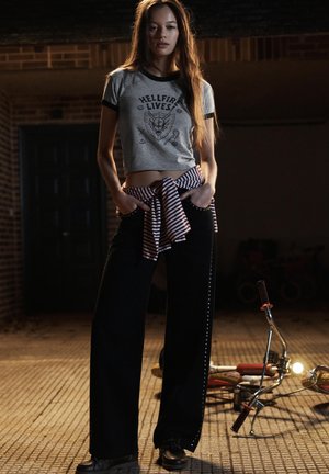 Gray cropped t-shirt with graphic print, black wide-leg pants with side studs, red and white striped tied waist detail, and black shoes.