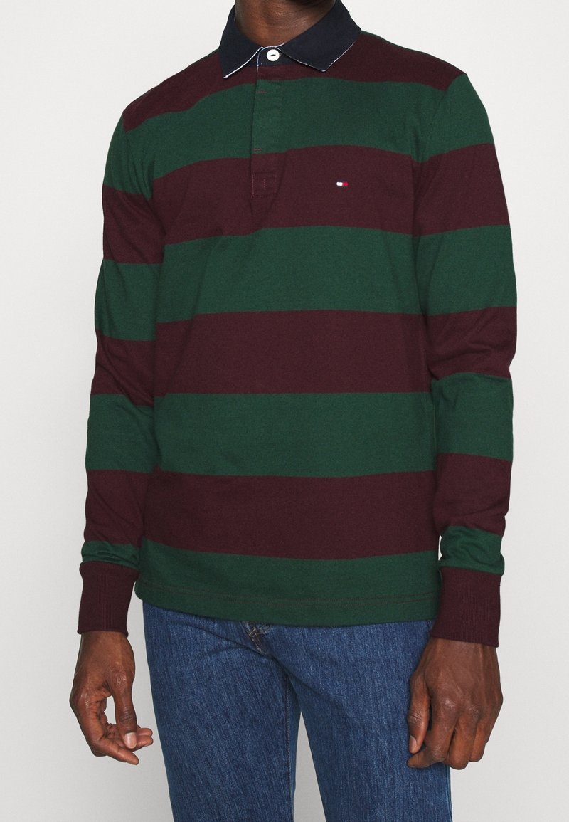 Homme portant un polo à manches longues avec des rayures horizontales vert foncé et bordeaux, un col noir et un jean bleu, debout devant un fond blanc.