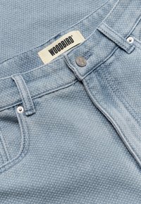 Jean en denim bleu clair texturé avec un bouton métallique et des rivets, passant de ceinture, et une étiquette en tissu portant la mention "WOODBIRD" cousue à l'intérieur de la ceinture.