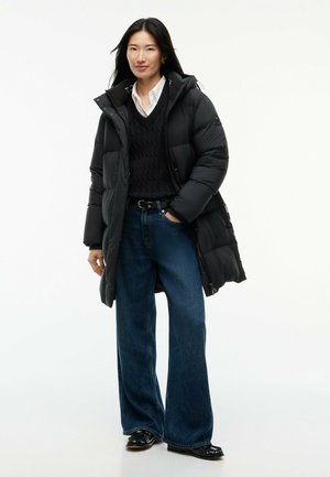 Kvinde iført en sort dunjakke over en sort sweater og hvid skjorte, brede blå jeans og sorte sko, stående med den ene hånd i lommen.