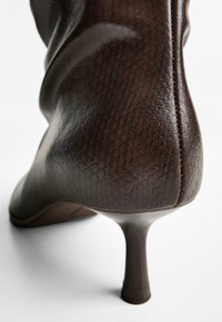 Botine din piele maro cu toc înalt, cu vârf ascuțit, suprafață texturată și toc stiletto subțire. Sămburi netezi și un luciu subtil sunt prezente.