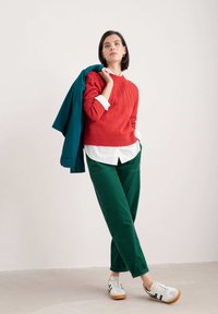 Röd kabelstickad tröja, vit skjorta med krage, gröna kordflorsbyxor, teal jacka liggande över axeln, och vita sneakers med gröna detaljer.