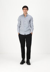 Chemise à boutons blanche et bleue avec un motif géométrique, associée à un pantalon noir et des chaussures noires, se tenant devant un fond uni.